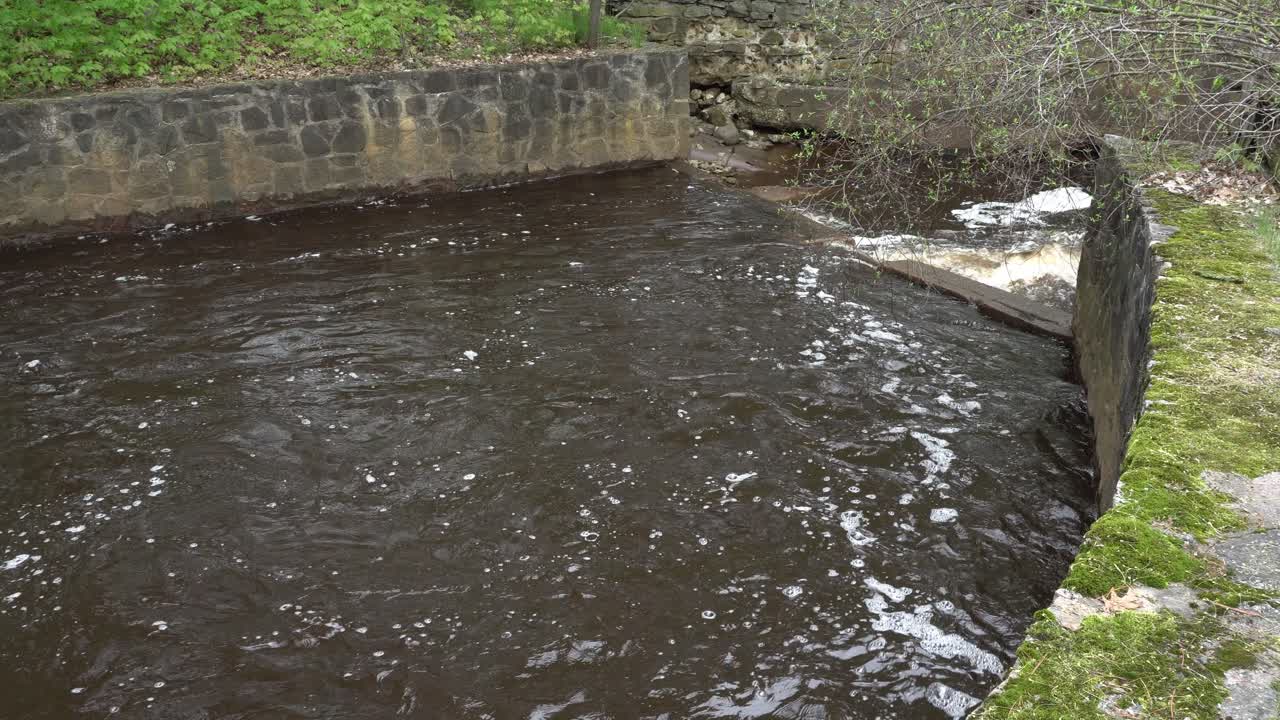 We see a beautiful view of a small river with stone walls on both sides. Additionally, there is some greenery on top of the stones, which makes it truly lovely.