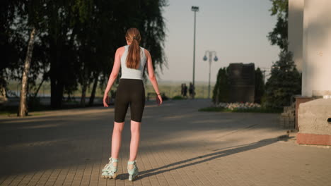 back view of young lady skating outdoors close to building on paved path, with blur view of people walking in distance, surrounded by lampposts, trees, and greenery in bright sunlight