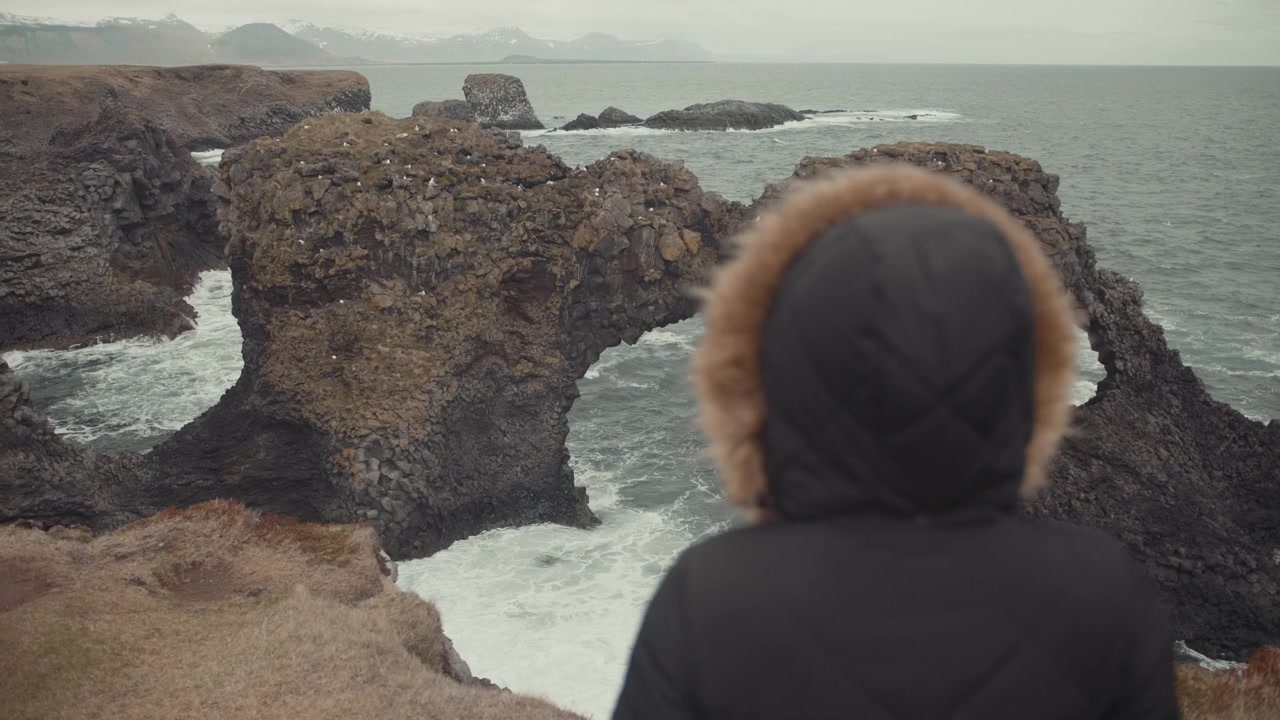 Person in Hooded Jacket Looking at Natural Rock Arch on the Coast