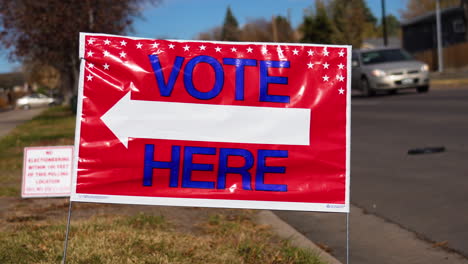 vote here sign pointing left with people driving cars in background, close up