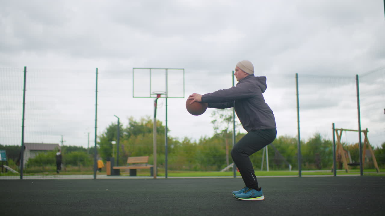 white senior man practicing basketball squat on urban community court, performing balance drills near hoop and fence with highrise backdrop, focused posture and rehab session energy, sneakers