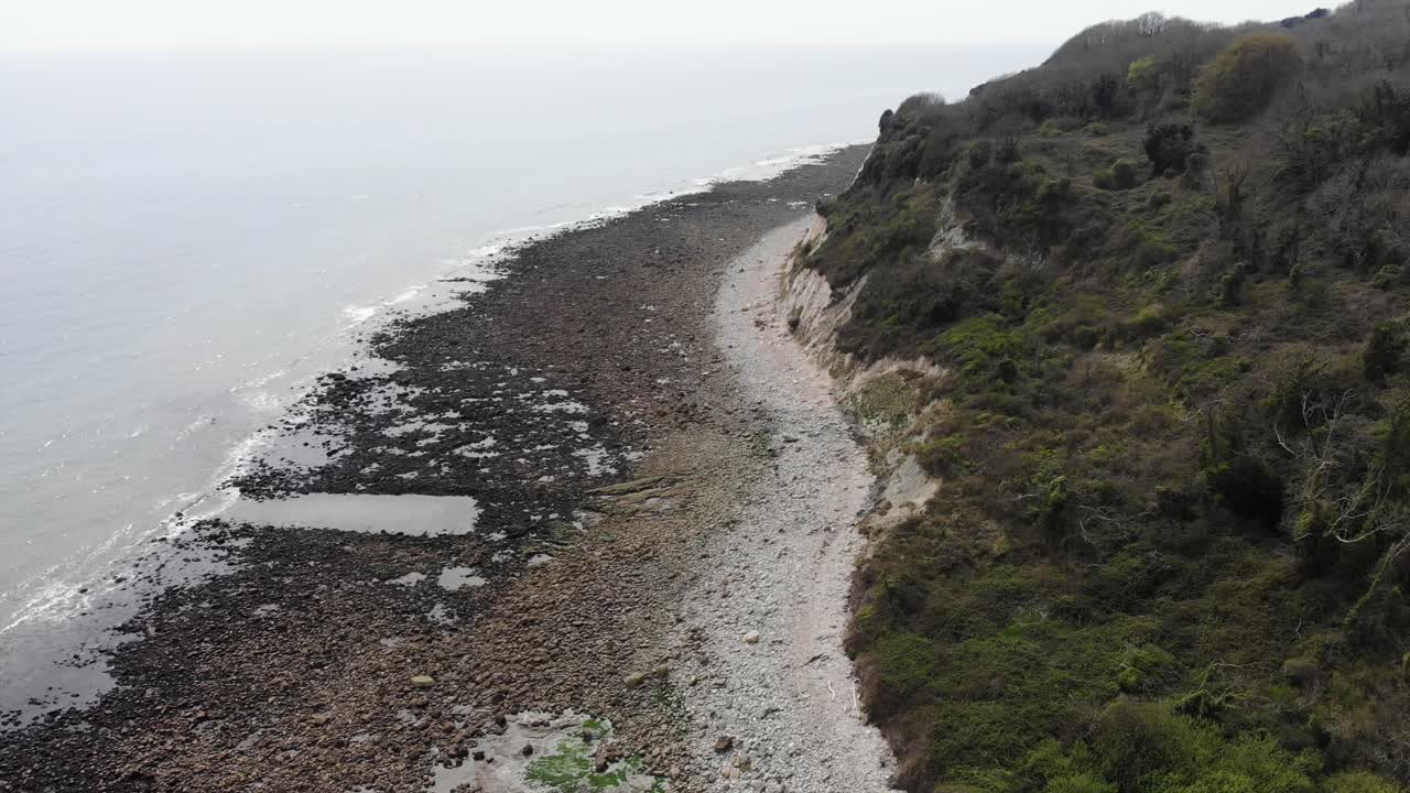 Aerial perspective showcasing the rugged coastline of Lyme Regis with Seaton undercliffs, highlighting the rocky beach and lush greenery. Dolly Forward Shot