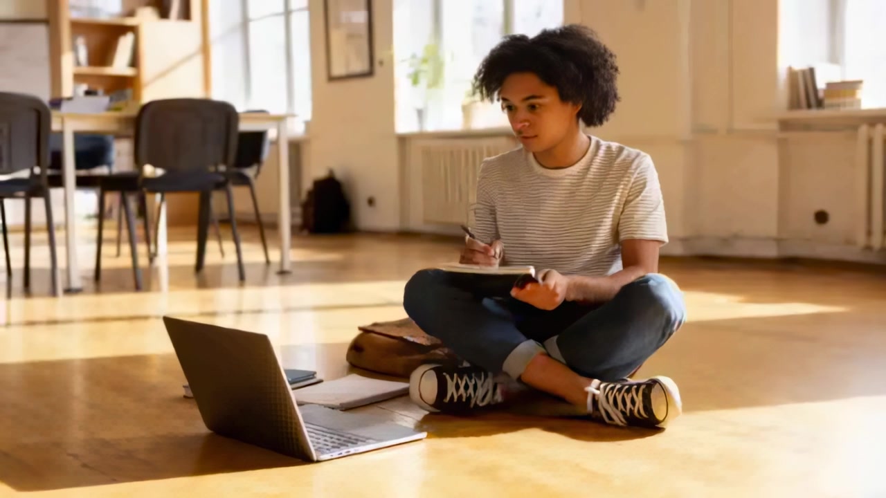 Turning, student in striped tee consulting laptop on floor, writing in notebook, studying