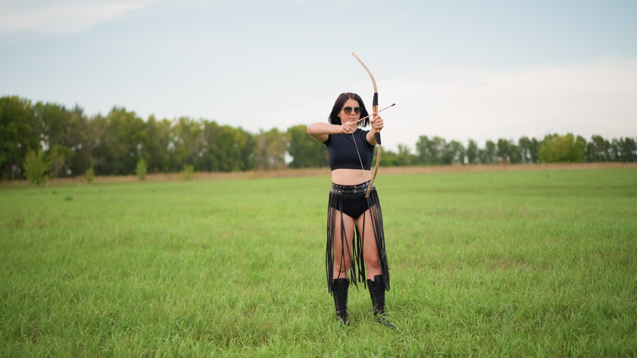 Mujer arquera apuntando en un campo de hierba, vestida con un traje de guerrera de cuero y botas, tensando un arco tradicional y soltando una flecha con una postura concentrada, pradera abierta y horizonte arbolado bajo un cielo suavemente nuboso