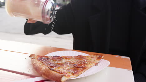 Woman-shakes-and-sprinkles-Parmesan-cheese-topping-on-pepperoni-pizza-slice-for-lunch-outdoors