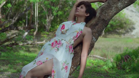 chica latina de pelo corto y rizado con un vestido largo yace en un tronco de árbol en un parque en un día soleado