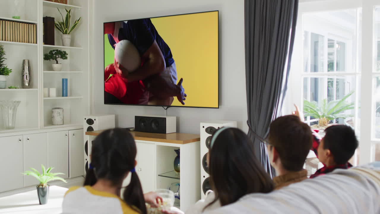 Asian family watching tv with diverse male rugby players playing on screen