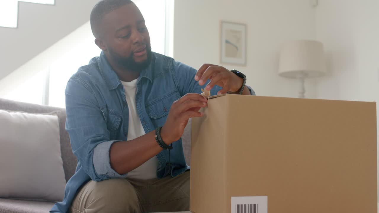 African American man inspecting tape, cutting with utility knife on box on sofa to retrieve order