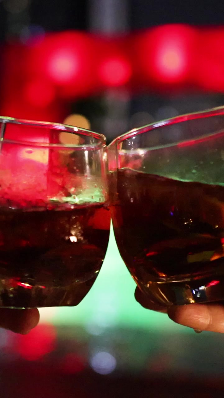 Two hands clink whiskey glasses with ice in a lively bar setting, colorful bokeh lights in the background, shallow depth of field, smooth camera movement