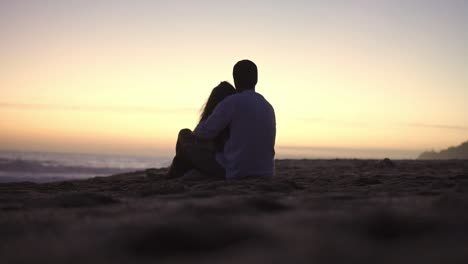 Static-handheld-shot-of-a-couple-in-love-in-silhouette-sitting-on-the-beach-in-front-of-the-sea-with-calm-waves-during-a-romantic-sunset