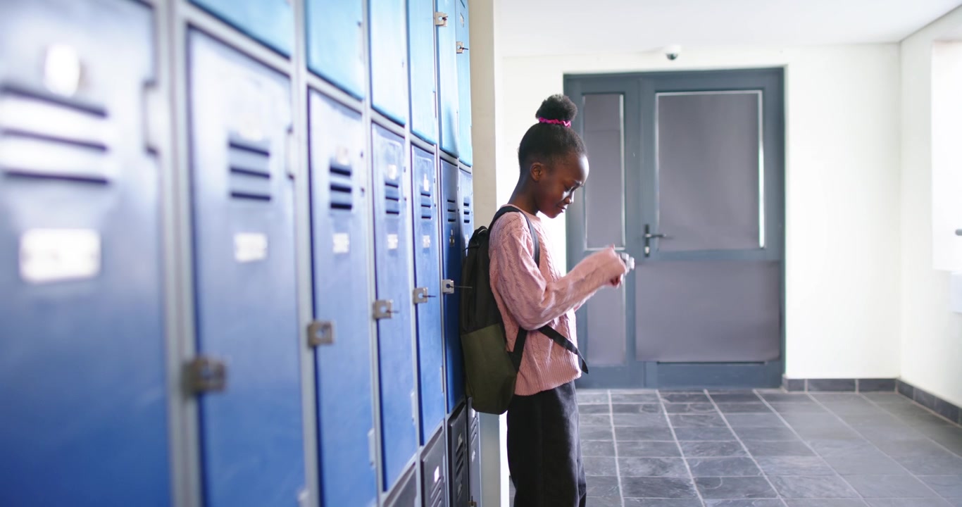 Three African American girls by lockers with backpacks, peers walking away, left girl covering face