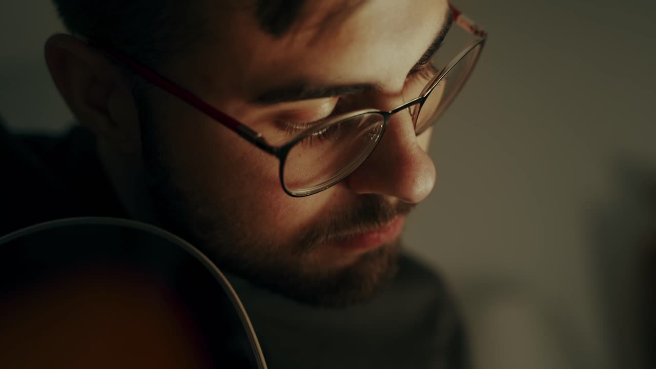 Face of a brunet male with beard that wears glasses looking at the neck of the guitar concentrated while he is playing