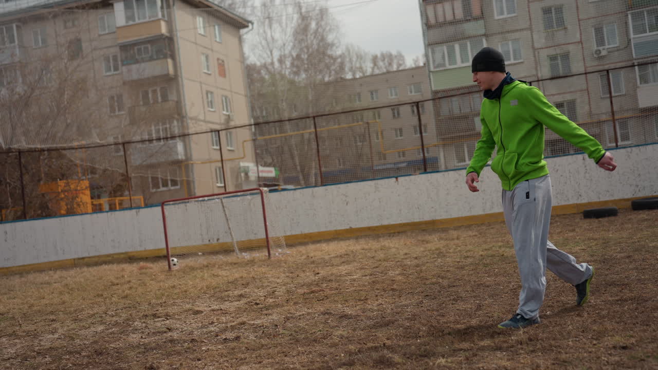 white man in bright green hoodie dribbles and volleys soccer ball across muddy urban court, beanie and grey sweatpants visible, aiming shots past small rusted goal with apartment backdrop