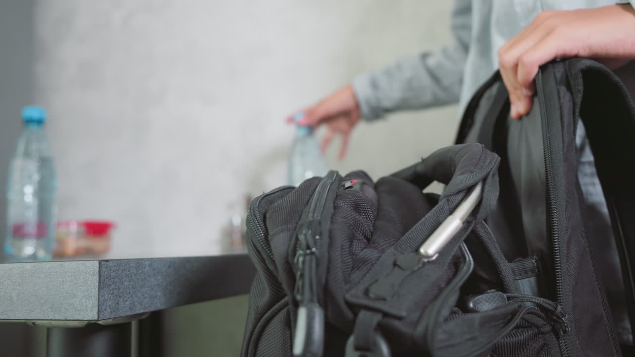 boy in casual shirt packing school bag on kitchen counter with notebooks, water bottles, lunch container, and pen holder in focus, organizing supplies before school day begins, preparing efficiently