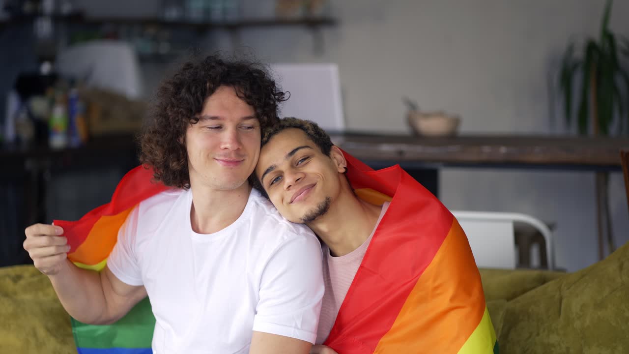 los chicos pasan tiempo juntos en casa en el sofá, se abrazan y acarician entre sí, bajo la colorida bandera del arco iris lgbt
