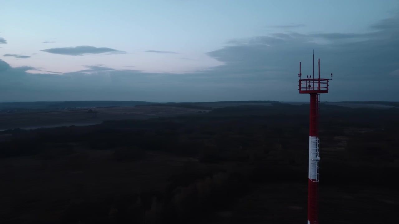 Vista aerea dall'alto al crepuscolo che mostra un'alta torre di comunicazione rossa e bianca che si erge accanto a una campagna aperta, con terreni agricoli scuri, edifici sparsi e una calma