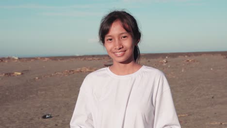 medium closeup of young female indonesian teen collecting litter from dirty polluted beach putting it in garbage bag then looking at camera smiling