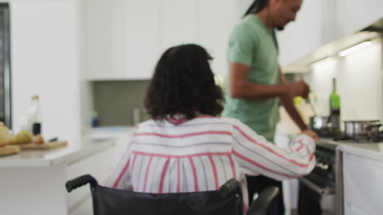mujer biracial en silla de ruedas hablando con un compañero de sexo masculino sonriente preparando comida en la cocina
