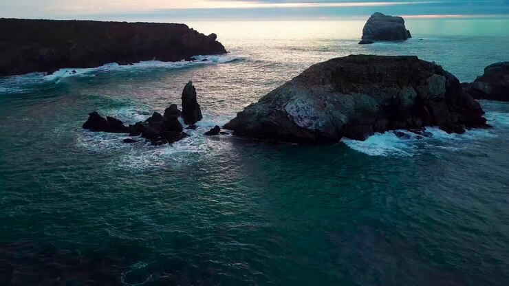 Volando tra enormi rocce oceaniche vicino a onde che si infrangono verso il sole che tramonta a Sand Dollar Beach a Big Sur, California.