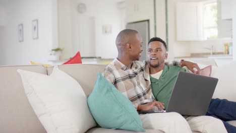 Happy-african-american-gay-male-couple-sitting-on-sofa,-embracing-and-using-laptop,-slow-motion
