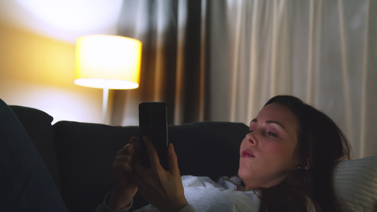 Woman Lying On Sofa At Home At Night Streaming Or Looking At Online Content On Mobile Phone 8