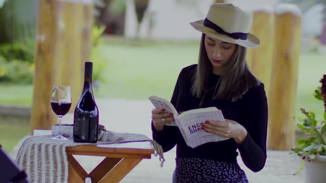 chica de moda con un libro de apertura fedora junto a una copa de vino tinto y una botella en una bodega