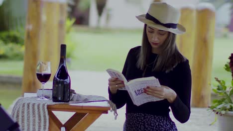 fashionable girl wearing fedora opening book by a glass of red wine and bottle at a winery