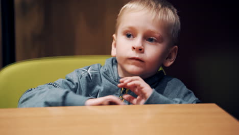 happy young boy sitting at the wooden table in the room. little blond kid with his arms on desk looking at camera. lovely child with a smile on his face.