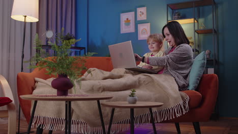 madre y niño pequeño hija niño estudio en la computadora portátil, viendo películas divertidas, relajándose, acostado en el sofá