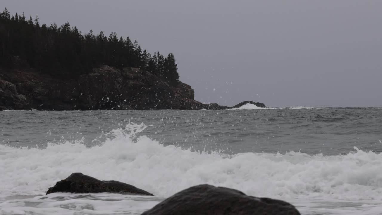 Rugged ocean waves break against rocks on shore under overcast gray sky, with dense forested cliffs in Cornwall UK, capturing moody coastal atmosphere and natural seascape