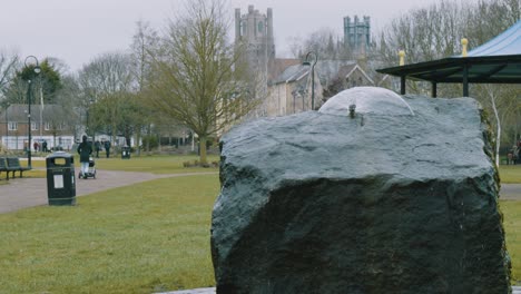 Mittlere-Aufnahme-Eines-Kleinen-Wasserbrunnens-In-Den-Jubiläumsgärten-Der-Stadt-Ely,-England,-Mit-Kathedralentürmen-Im-Hintergrund,-Zeitlupe