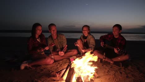 Picnic-with-friend-on-the-beach-near-the-bonfire.Young-people-are-roasting-sausages-on-wooden-sticks.-Male-from-the-side-is-playing-the-guitar.-Night-time