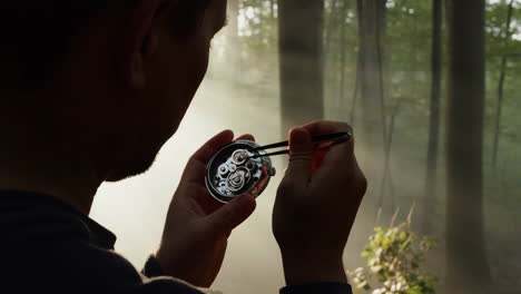 a person delicately working on a mechanical watch movement in a misty, sunlit forest