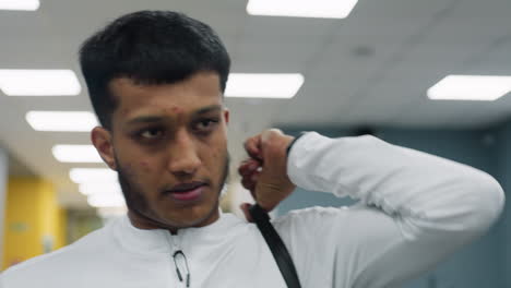 close up of student in white sportswear adjusting heavy black bag strap on shoulder while walking through gym corridor, with bright overhead lighting illuminating face and background
