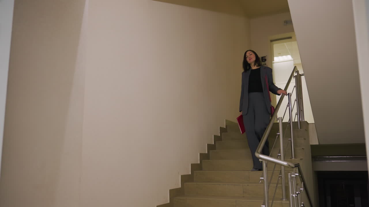Businesswoman walking up stairs in office building, carrying red handbag and folder. Confident and stylish in office attire, focused and ready for work in professional setting