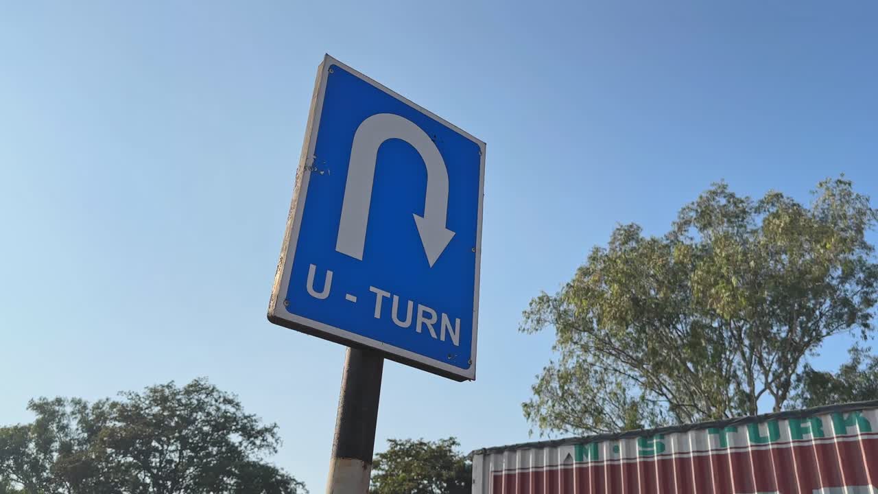 Tiefer Winkel, Aufnahme um ein blaues U-Turn-Verkehrsschild mit gebogenem Pfeilsymbol, vor klarem Himmel und Bäumen am Straßenrand, Sonnenlicht erzeugt sanfte Highlights auf dem Schild