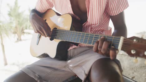 Midsection-of-african-american-man-sitting-playing-guitar-on-sunny-beach,-slow-motion