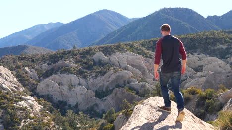 a young man on a hike walking out and sits down in slow motion on the edge of an amazing mountain peak in the california forest