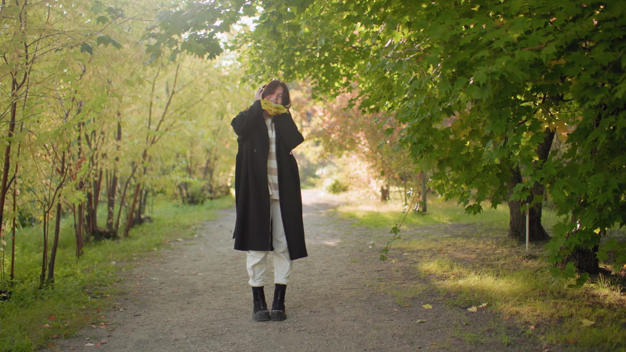 Nature explorer standing with bunch of yellow leaves in mouth while wearing headphones, moving body in excitement under bright sunlight, enjoying playful moment during autumn walk