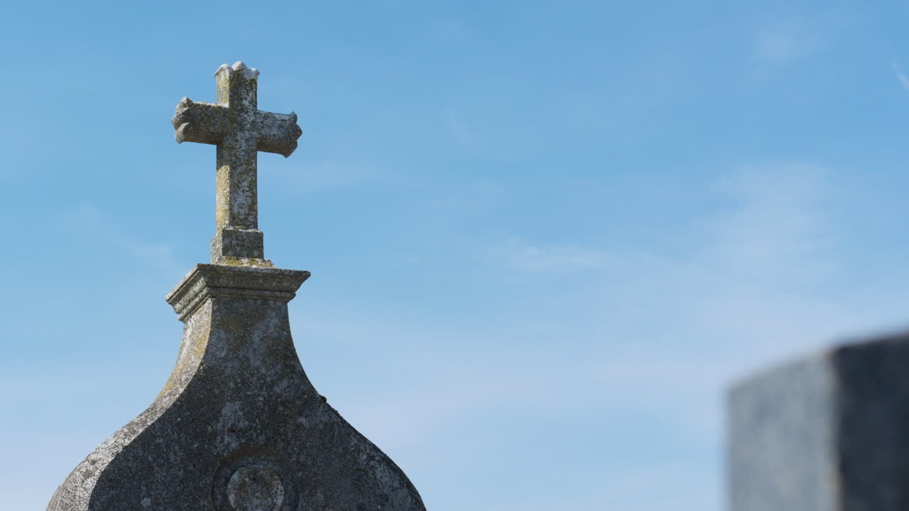 Weathered stone cross atop a tombstone in a cemetery, set against a serene blue sky