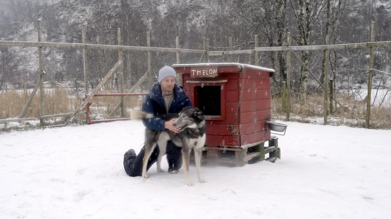 Mand knæler i sneen ved siden af et lille hundehus, mens flere hunde løber hen for at hilse på ham, modtage klap og kærlighed.
