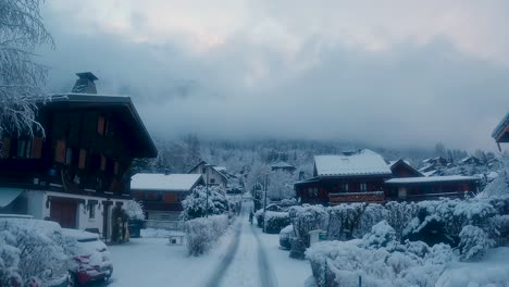 Man-carrying-skis-in-snowy-village-with-huts,-snowy-bushes,-and-cloudy-sky