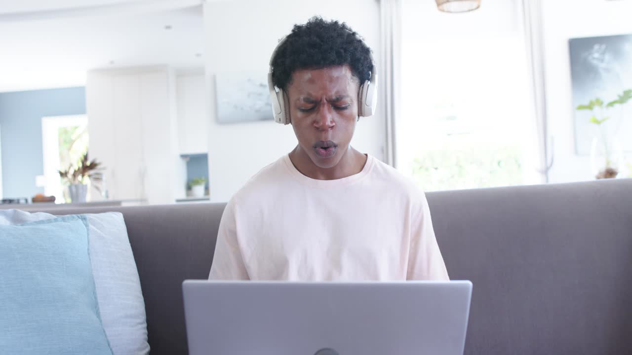 Uomo afroamericano ascolta una canzone sul laptop, indossa le cuffie e canta in silenzio, poi sorride sul divano.
