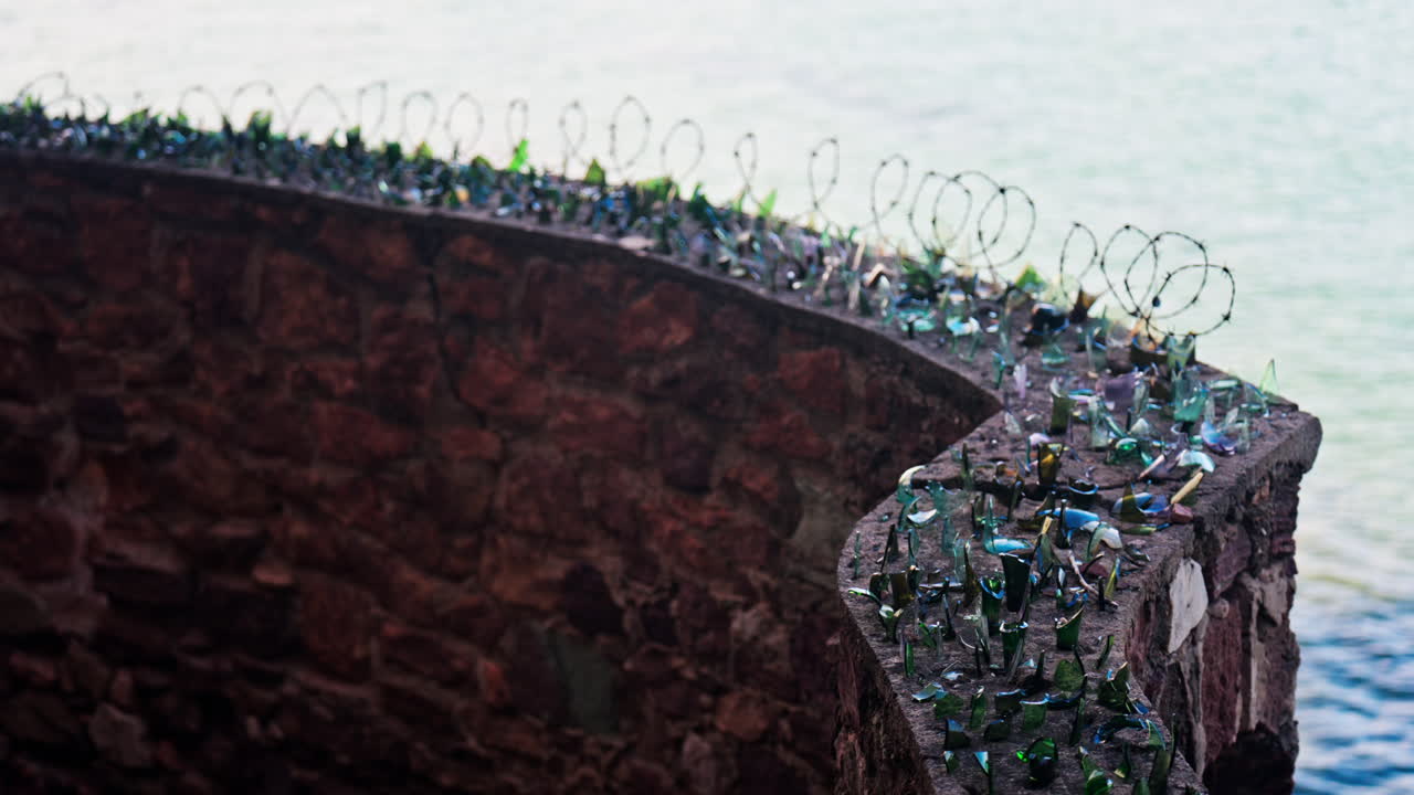 Close up of multiple broken glass shards and barbed wire loops along the edge of a brick wall with a blurred view of the sea