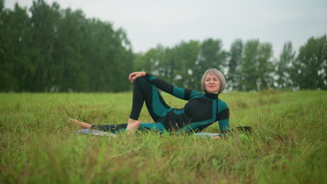 middle aged woman lying on her side with hand resting on her leg, looking thoughtfully into the distance, surrounded by greenery and trees in the background