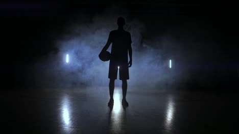 Die-Silhouette-Eines-Basketballspielers-Auf-Dunklem-Hintergrund-Mit-Rauch-Auf-Dem-Basketballplatz-Wirft-Einen-Basketball-Und-Blickt-In-Zeitlupe-In-Die-Kamera