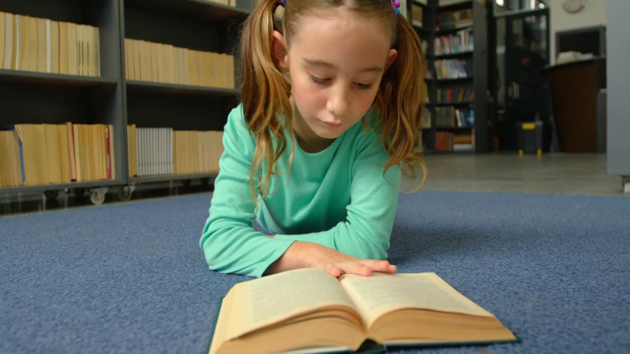 Vista frontale di una scolaretta caucasica attenta che legge un libro in biblioteca a scuola 4k