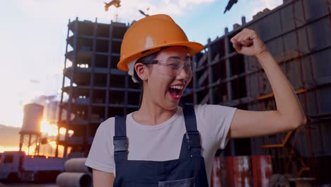 close up de una trabajadora asiática que usa gafas de protección y casco de seguridad sonriendo y flexionando su bíceps mientras está de pie en el sitio de construcción