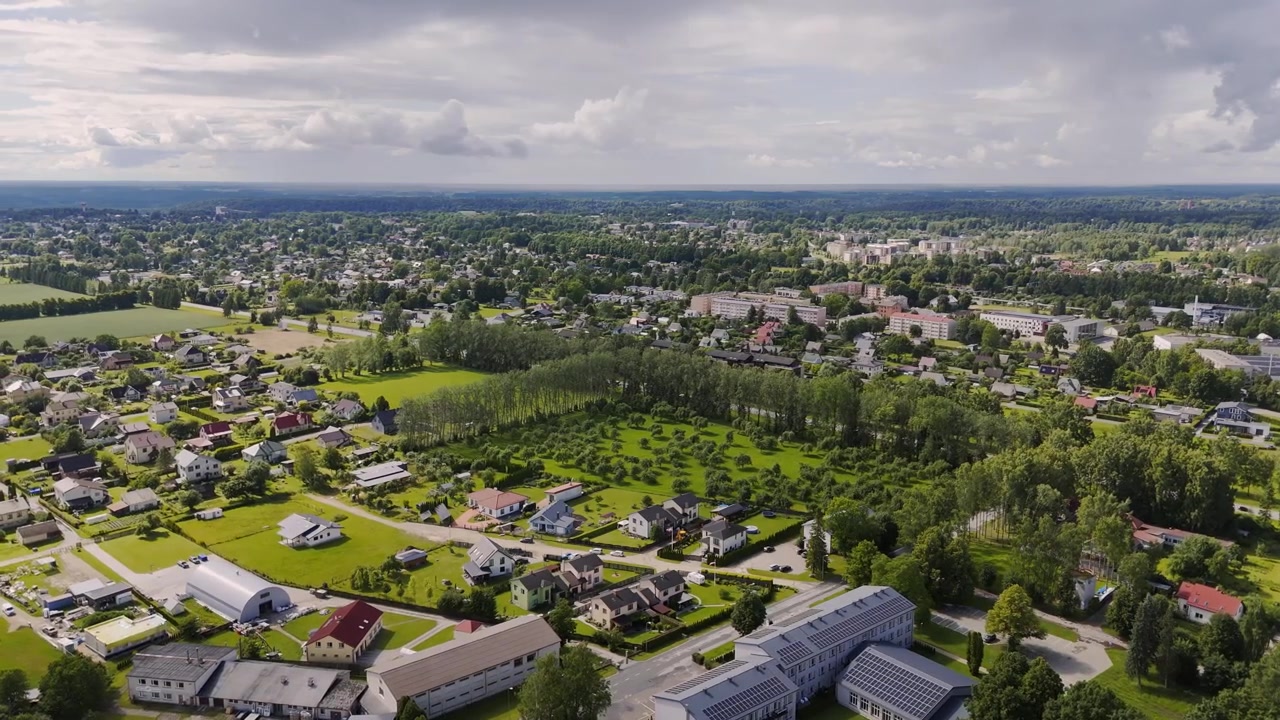 Aerial drone view over Sigulda, Latvia, showing residential homes, green fields, tree-lined neighborhoods, solar panel rooftops and distant townscape under cloudy sky.