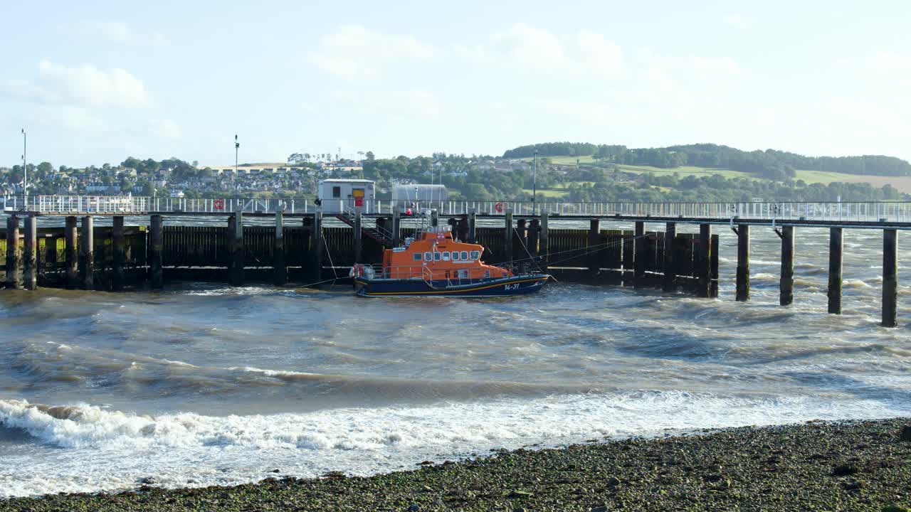 An orange lifeboat navigates choppy waves, approaching a pier at Broughty Ferry, Scotland